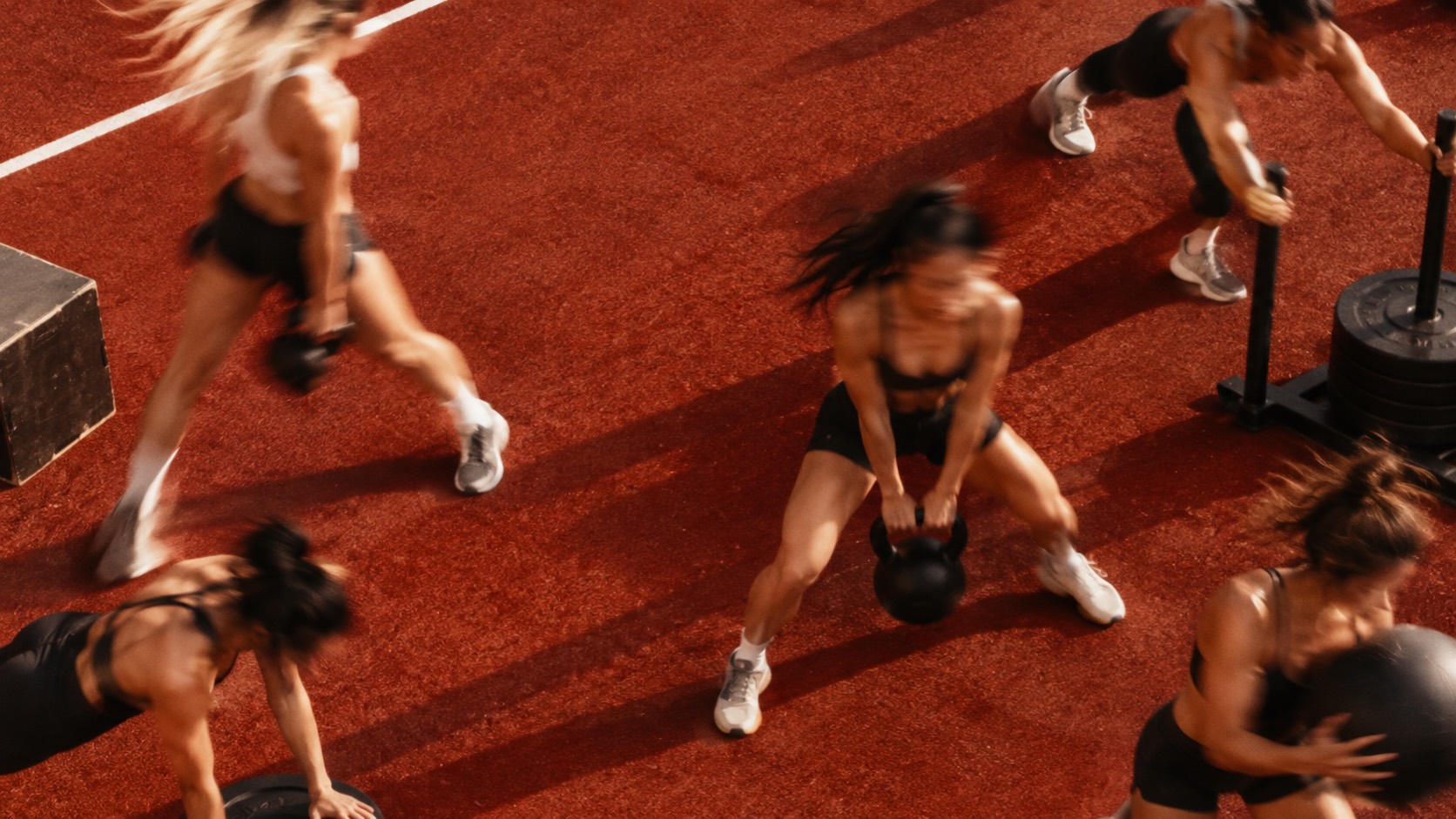 Women training on a red track: a kettlebell swing, sled push, box step and medicine ball, blurred with movement